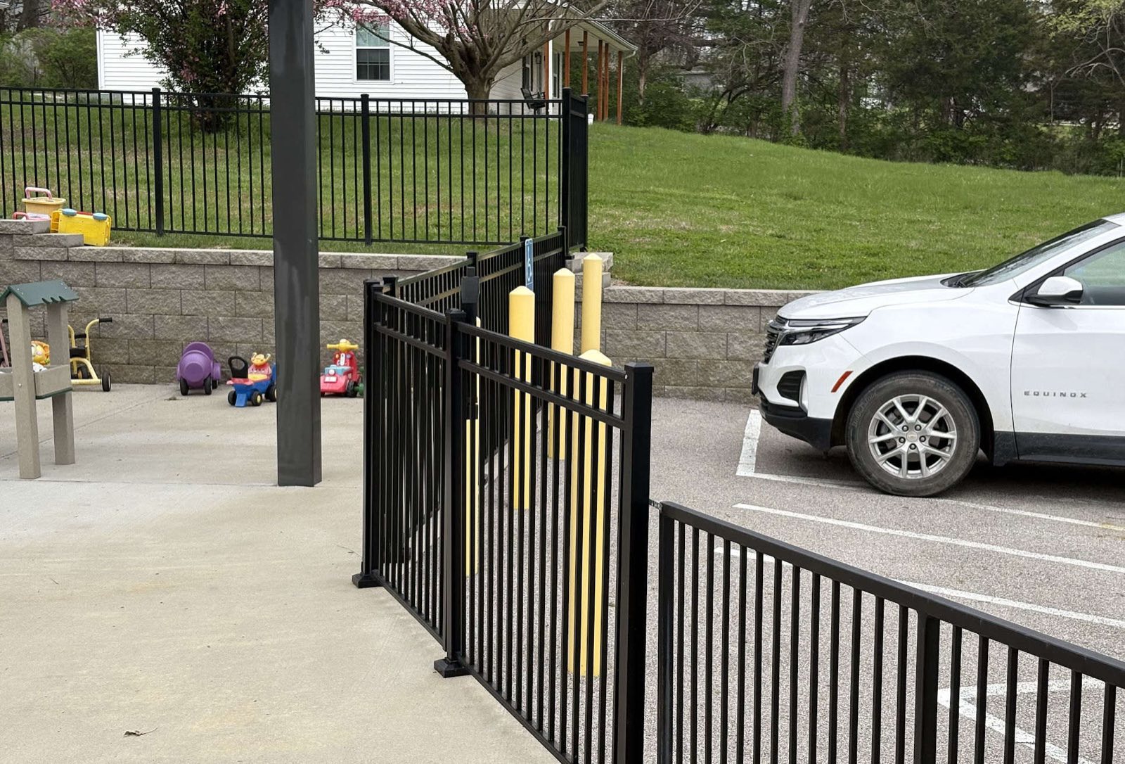 Ride-on toys in a fenced play area at an early childhood program, with yellow bollards and a fence standing between the children's space and a car parked a few feet away